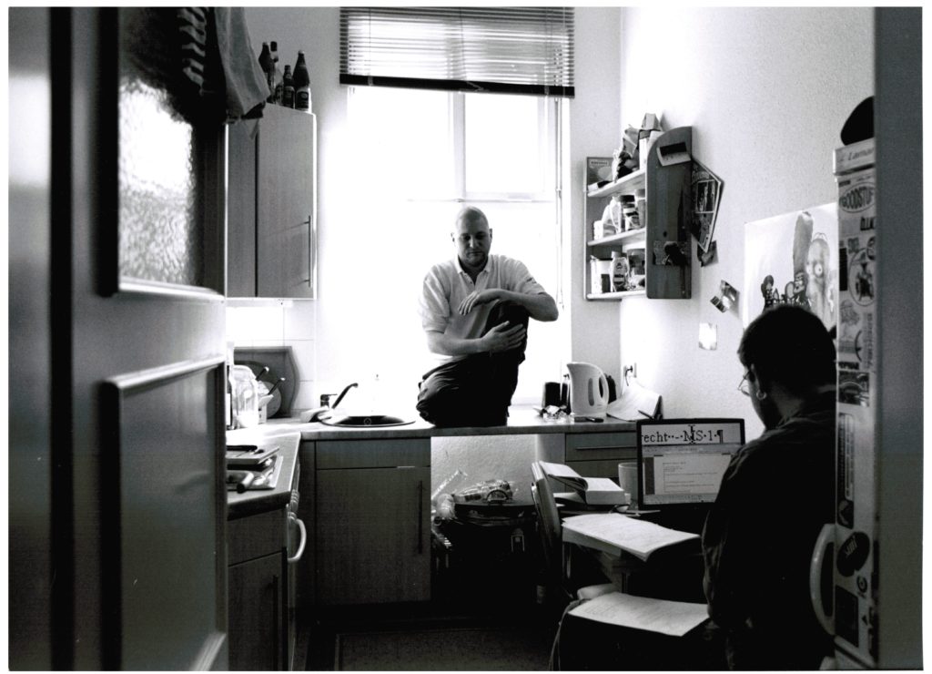 Photo Carsten 2009 - Carsten sits with his study assistant in a kitchen. The study assistant is sitting in front of a computer and is sorting paper documents.
