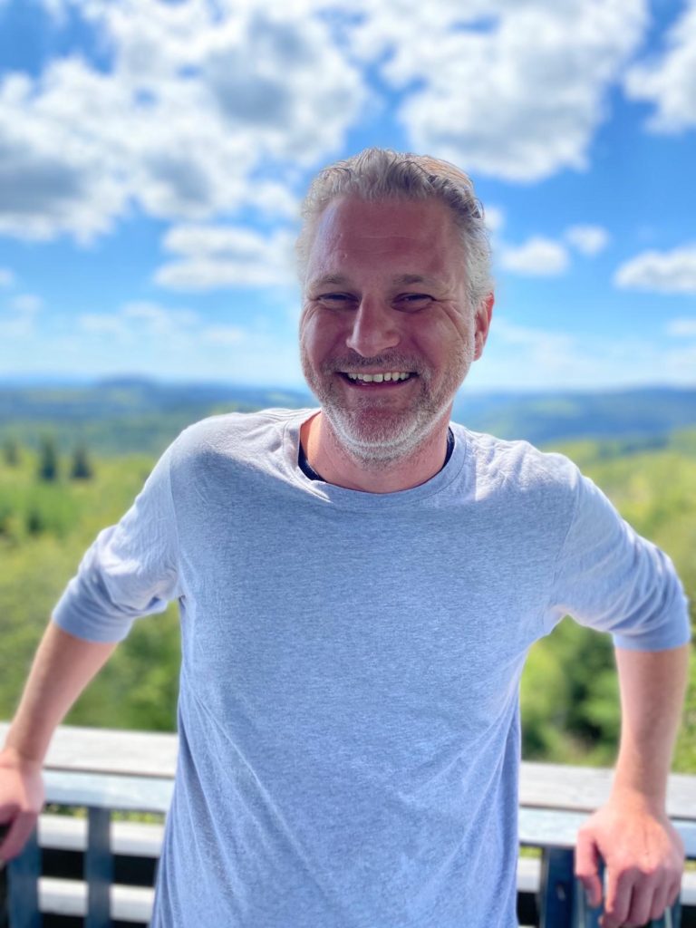 Portrait of Carsten Otto - a laughing man with gray curly hair and three days beard in front of a blue sky with clouds.
